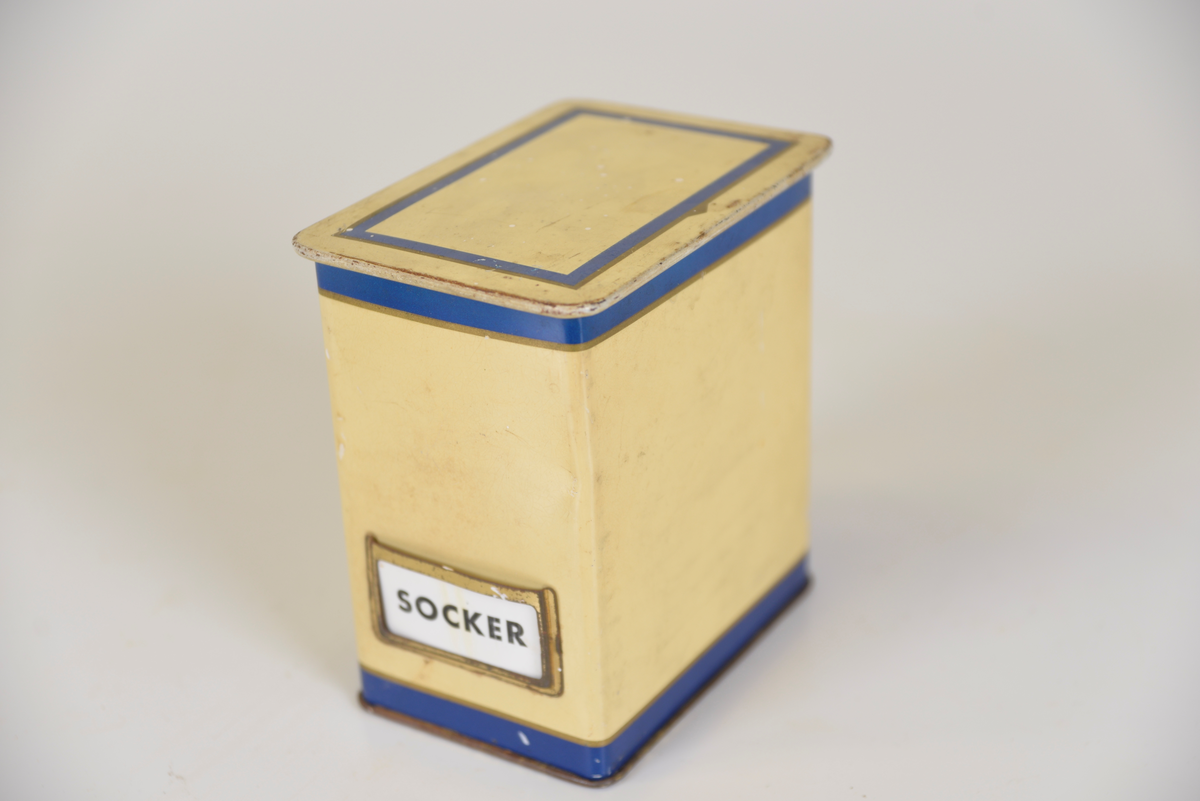 Gula ålåtburkar med blå dekor. Etiketthållare , två med KAFFE tryckt på burken, en med vit plastetikett med text "SOCKER" i svart och en med etikett med "KAKAO".  Det kan vara så att när man köpt kaffe har man fått det i burken samt lösa etiketter för att återanvända burken till annat när kaffet var slut.

Från Arne Ekströms handelsmuseum.