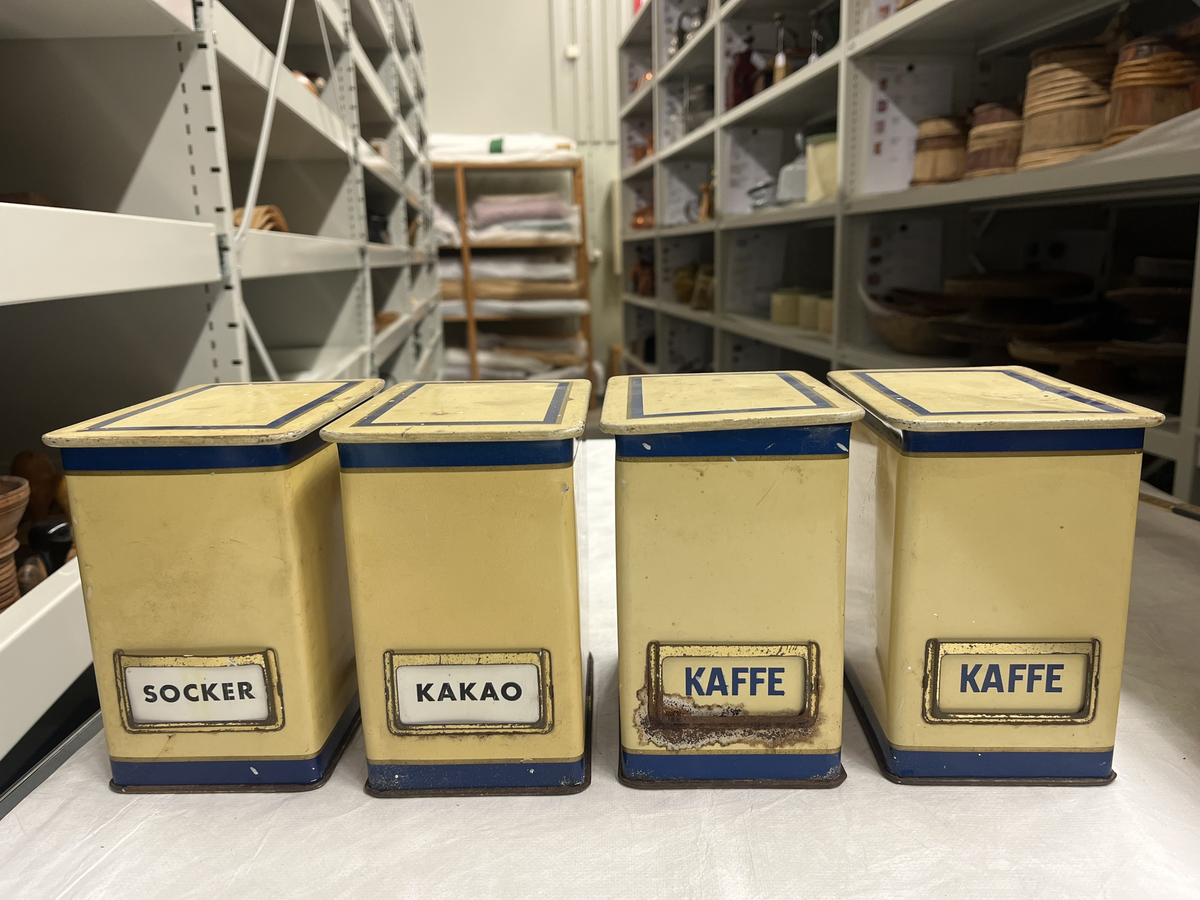 Gula ålåtburkar med blå dekor. Etiketthållare , två med KAFFE tryckt på burken, en med vit plastetikett med text "SOCKER" i svart och en med etikett med "KAKAO".  Det kan vara så att när man köpt kaffe har man fått det i burken samt lösa etiketter för att återanvända burken till annat när kaffet var slut.

Från Arne Ekströms handelsmuseum.