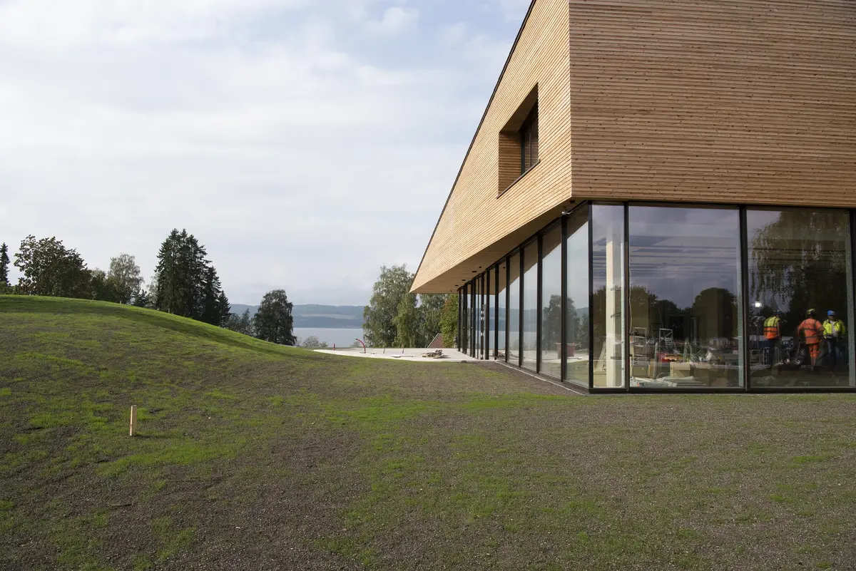 Nysådd bakke som bølger ned mot det regionale historiesenteret med sin trekantkonstruksjon i treverk over store glassflater med speilbilde av arbeidere i signalklær.