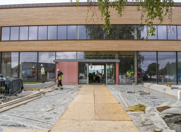 Fasadebilde med fine linjer; lyst, liggende treverk i en bred stripe øverst, deretter vinduer i "normal" høyde før ny, smalere treverksstripe og store vindusflater. Midt på: inngangsdør i glass, med delvis tildekking. Betongen utenfor er dekket av hvit markduk, treplater er lagt oppå for å gjøre det mulig å gå inn enn så lenge.