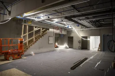 En oransje lift står i ene hjørnet, taket er åpent inn til teknisk lag med mange rør og ledninger, byggematter dekker golvet, en trapp kommer ned langs ene veggen.