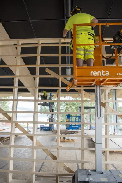En snekker i signalklær står høyt i en lift og bygger en lettveggkonstruksjon.