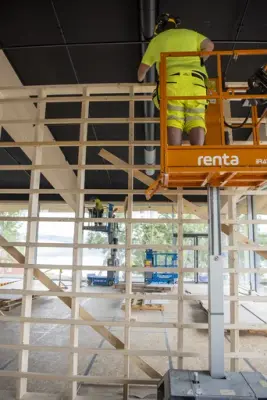 En snekker i signalklær står høyt i en lift og bygger en lettveggkonstruksjon.