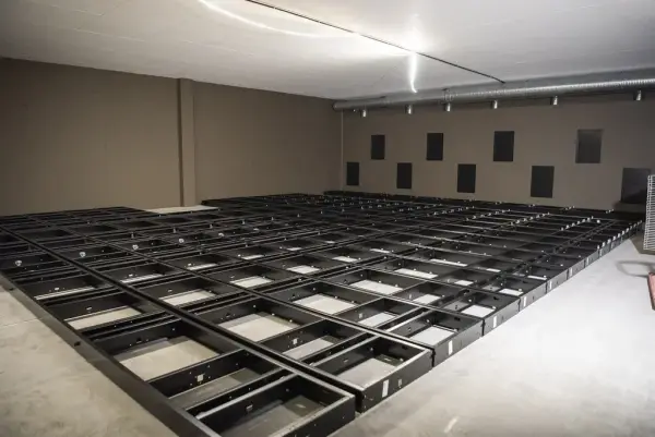 Lysegrå vegger, den ene med mørkere firkanter i tannmønster. På golvet ligger bunnrammene til arkivreoler som et mørkt firkantmønster.