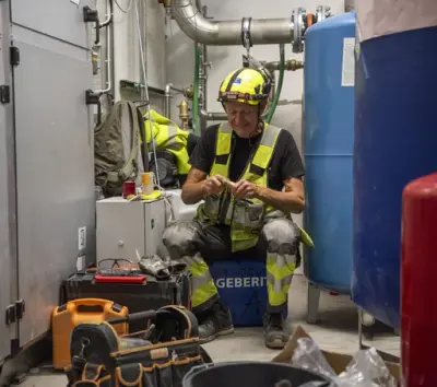 Mann i varselklær sitter på en blå kasse med verktøykassa foran seg og bruker fingrene på detaljarbeid. Rundt ham er rør, grå skap og blå tank.