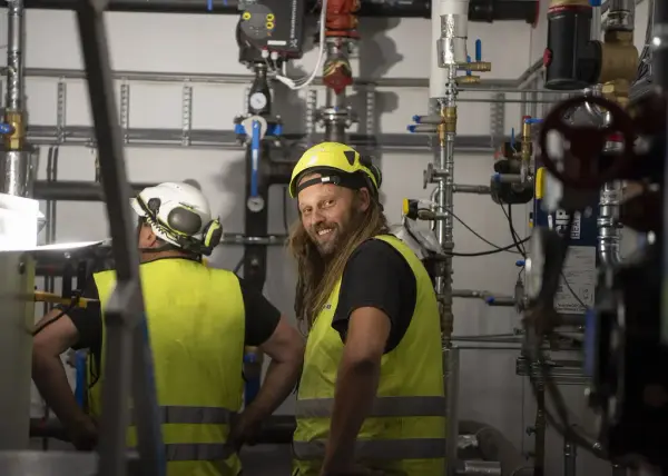 To rørleggere i signalklær står blant masse rør og koblinger i det regionale historiesenteret, den ene rørleggeren snur seg mot kameraet og smiler godt.