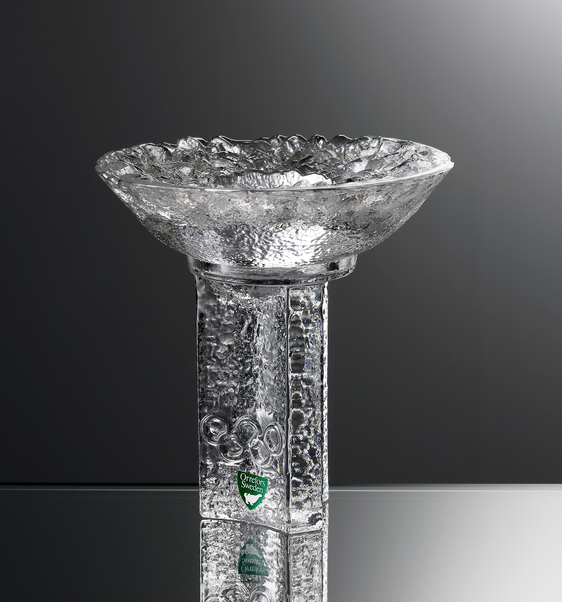 Glasskulptur i form av en OS-fackla, formgiven av Lars Hellsten 1985. 
Grön etikett med en vit orre (vilket markerar sekundaglas).