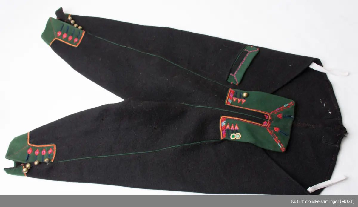 Bukse har fire hovedstykker i vadmel. Midtsøm framme og søm fra låket har innlagt grønn titte kant.
Den indre sømmen på bukse beinet går et stykke opp i baken før den møter baksømmen.
Bukse har smalt låk, belagt med grønnt klede med brodere og åtte knapphull og tre knapper. Tre knapper mangler. En kuleknapp i messing. To knapper i messing og grønn glass.
Lommeklaff oppstående (høyre side) belagt med grønnt klede med broderi. Grønn titte kant nede.
Lommefor lerret et firkantet stykke på sydd som lomme.
Nede, buksebein smale nederest bredde. Kantet med grønnt klede og opp splitt. Fire knapphull, fire messing kuleknapp og ullbroderi.