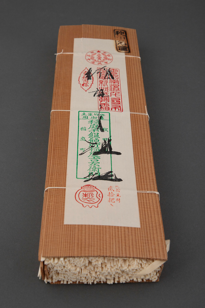 Pakning laget av to tynne plater i japanseder, med smalere plater på sidene, ombundet med tre tynne snorer. På den ene platen er det et svimerke og en påklistret papiretikett. I pakningen ligger tørkede nudler ombundet med tre bånd.
