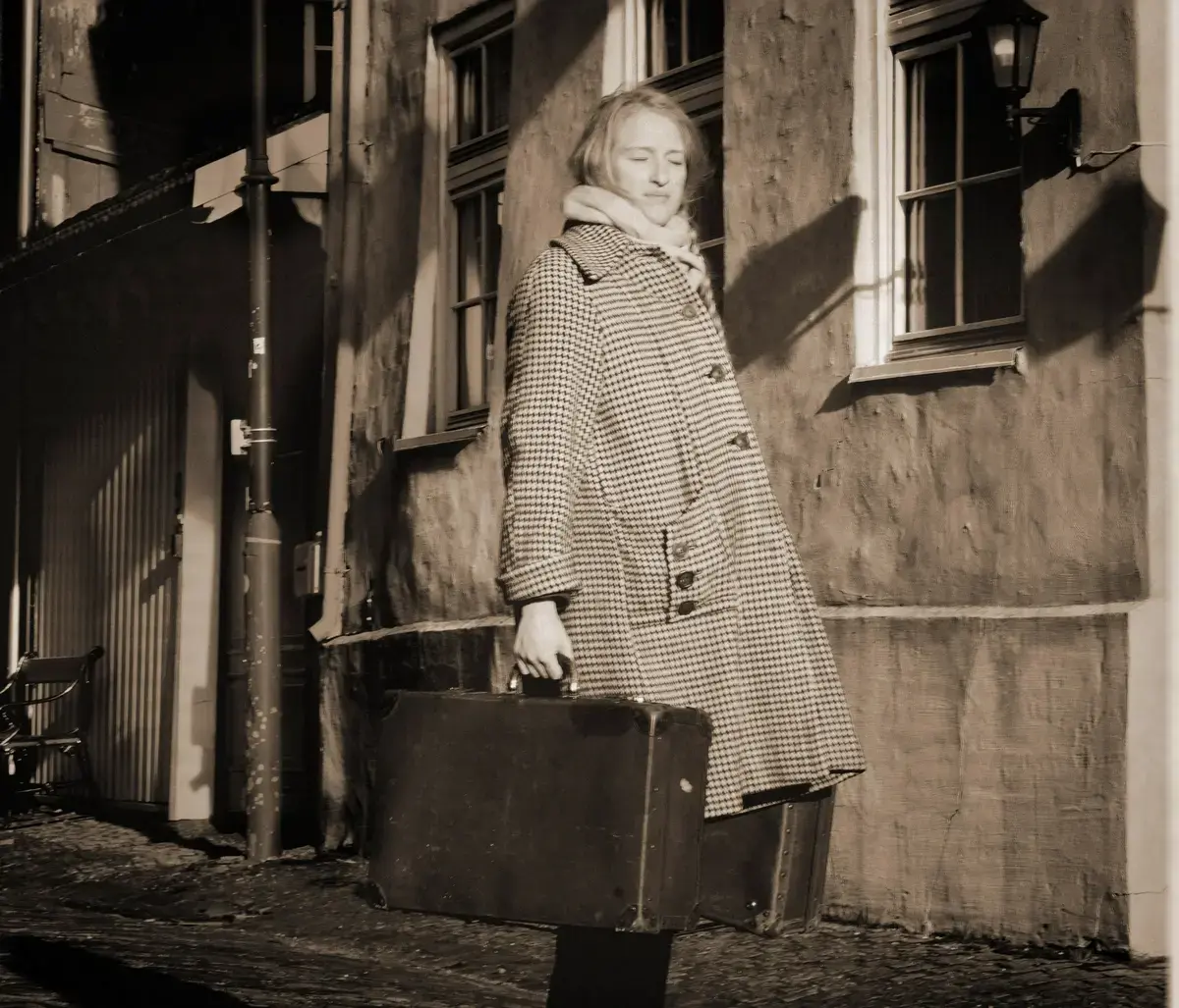 Svart/kvitt fotografi av ei kvinne som står ute på gata foran et murbygg. Kvinna har på seg ei kåpe og har en koffert i hver hånd, øynene er lukket.