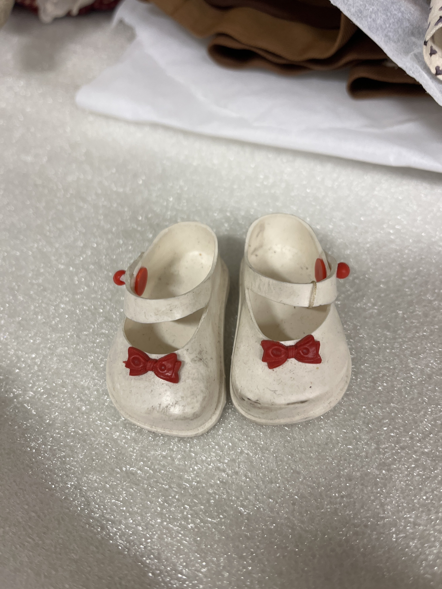 Huvud och armar av skinn, peruk av blont hår
Klädd i rutig klänning i rött och vitt samt hatt av flätad halm
(Strumpor och skor från 1950-t.)
Till dockan hör:
2 st underkjolar av vit bomull
Kappa av brun plysch
Randig bomullsklänning
Prickig bomullsklänning
1 st linne

Dockan har tillhört givaren mor
Funktion: leksak