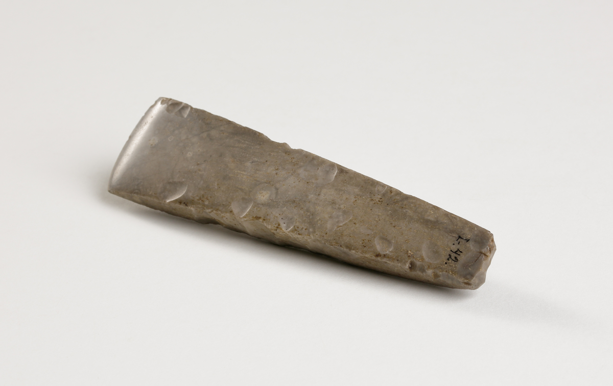 Flintmejsel av grå flinta, närmast tunnackig, slipad med avrundad egg. Rektangulär genomskärning.
