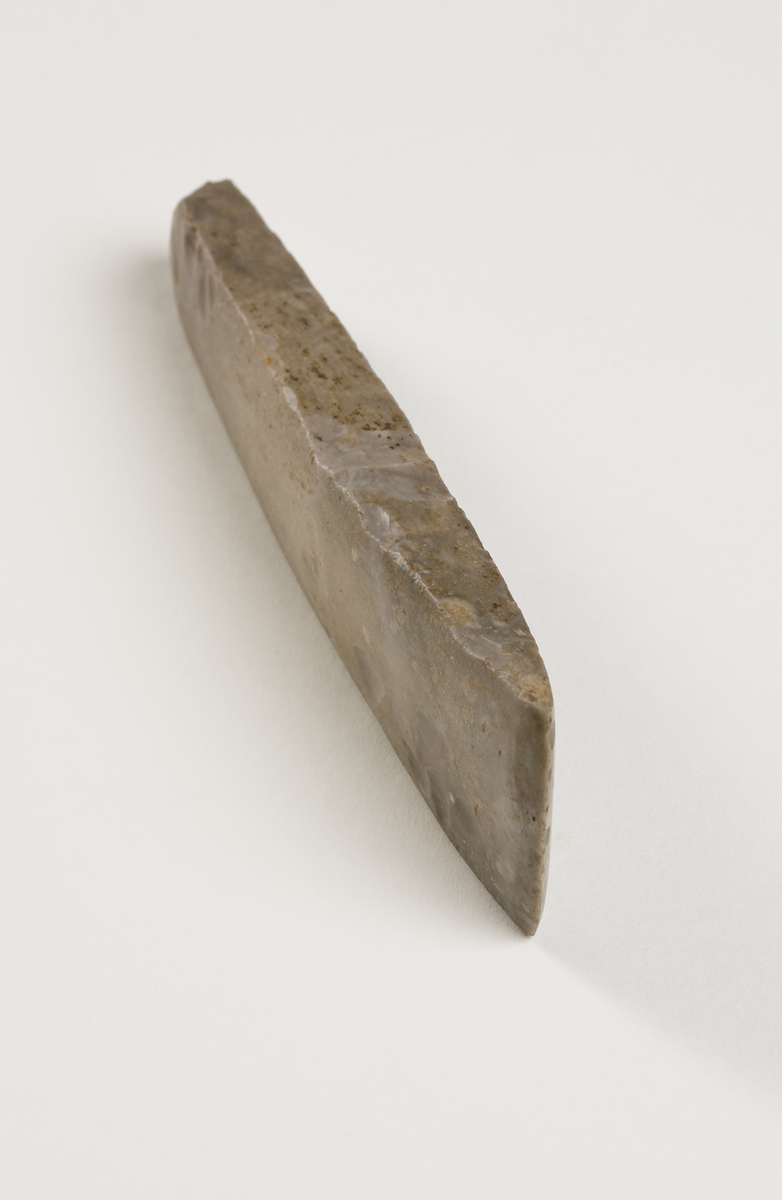 Flintmejsel av grå flinta, närmast tunnackig, slipad med avrundad egg. Rektangulär genomskärning.