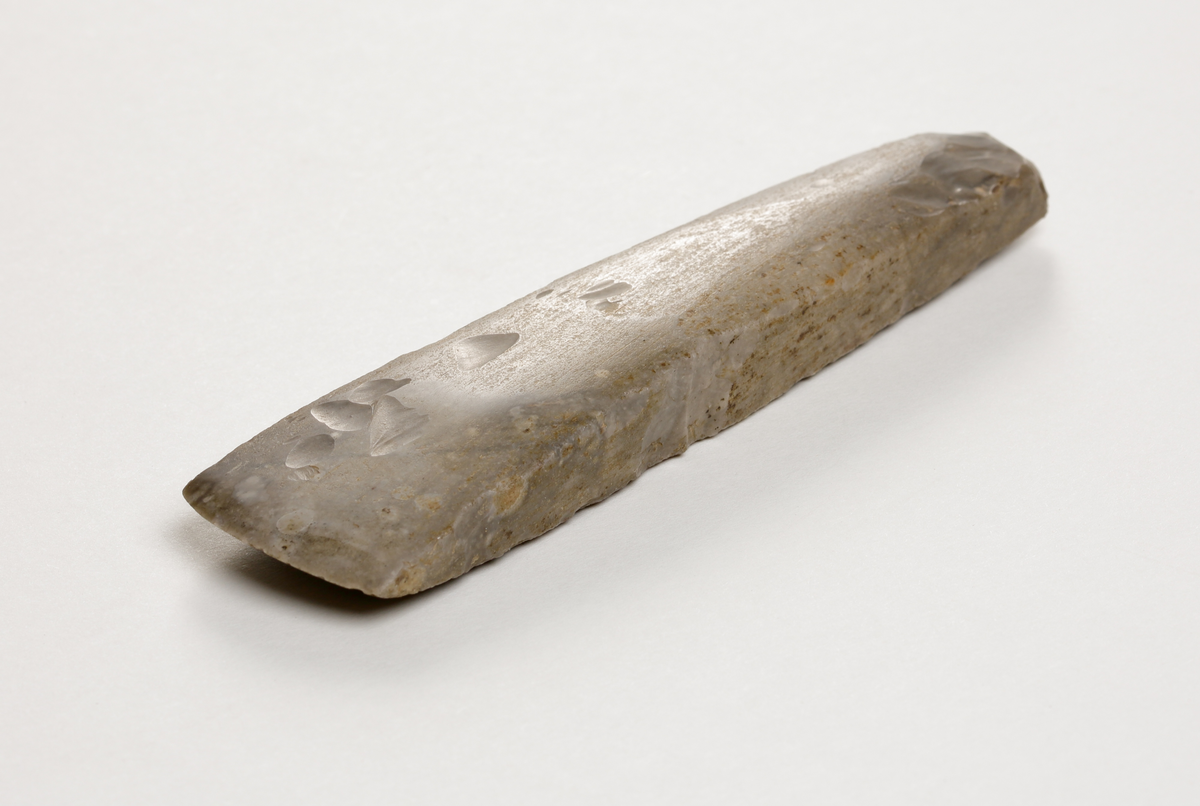 Flintmejsel av grå flinta, närmast tunnackig, slipad med avrundad egg. Rektangulär genomskärning.