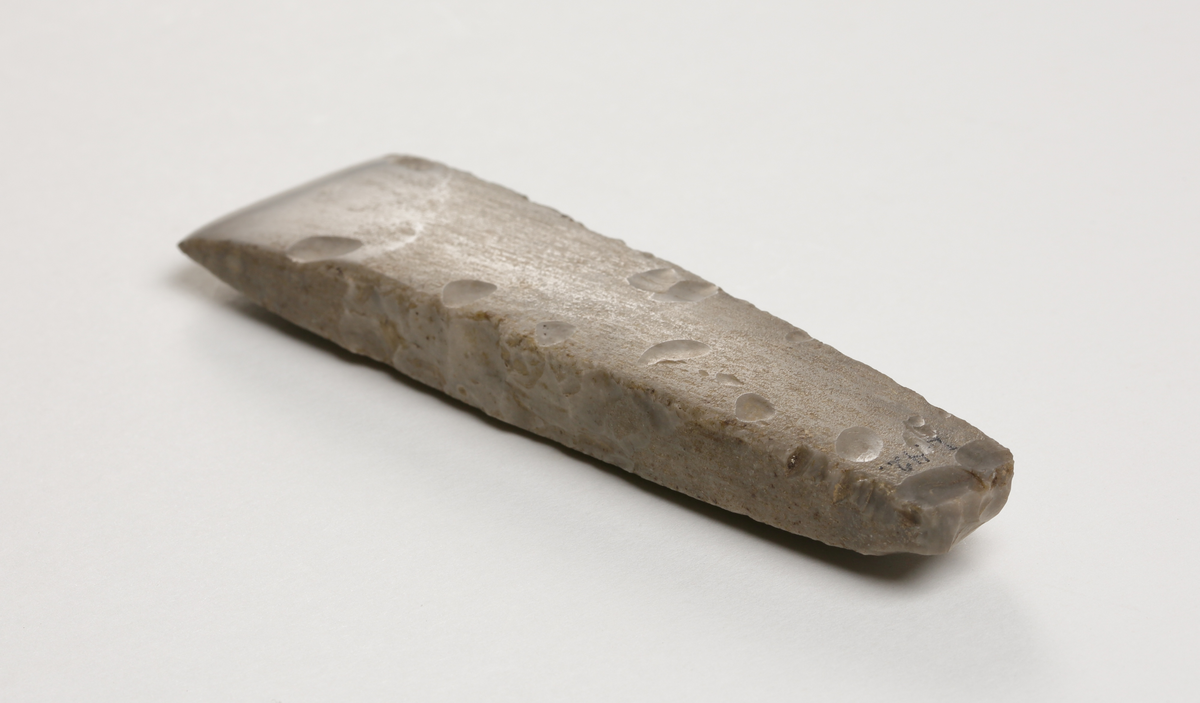 Flintmejsel av grå flinta, närmast tunnackig, slipad med avrundad egg. Rektangulär genomskärning.