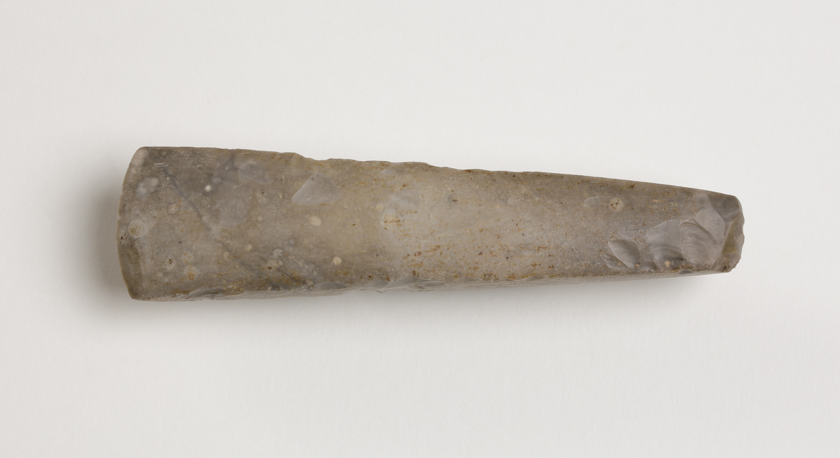 Flintmejsel av grå flinta, närmast tunnackig, slipad med avrundad egg. Rektangulär genomskärning.