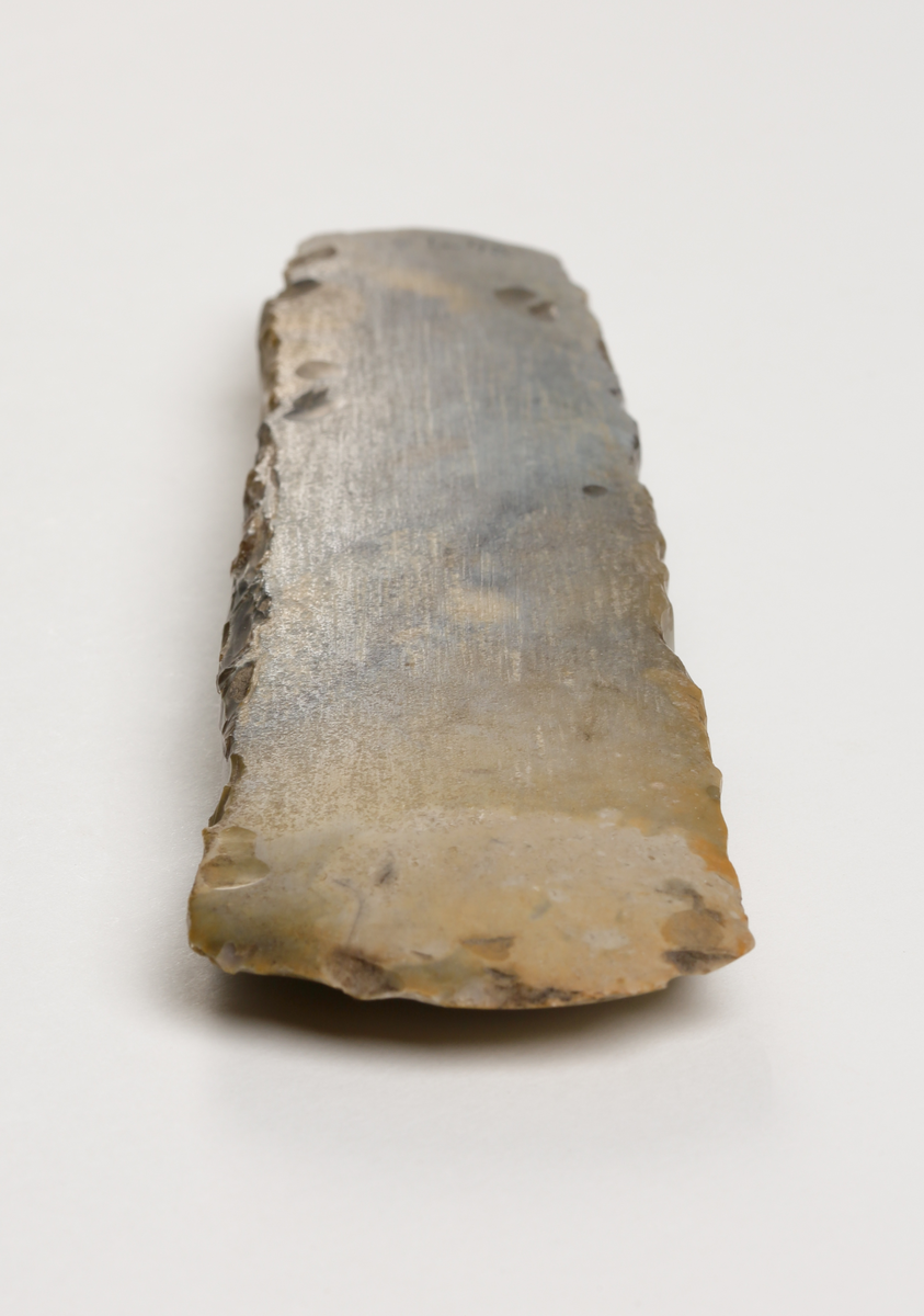 Flintyxa av brungrå flinta, närmast tunnackig, slipade bredsidor med avrundad egg. Rektangulär genomskärning.