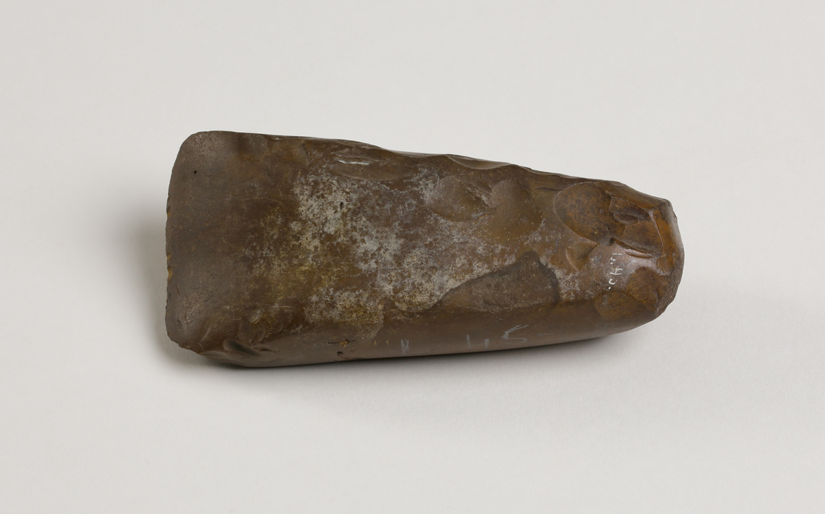 Flintyxa av brun flinta, tjocknackig, slipad och svallad, svagt håleggad. Kvadratisk genomskärning.