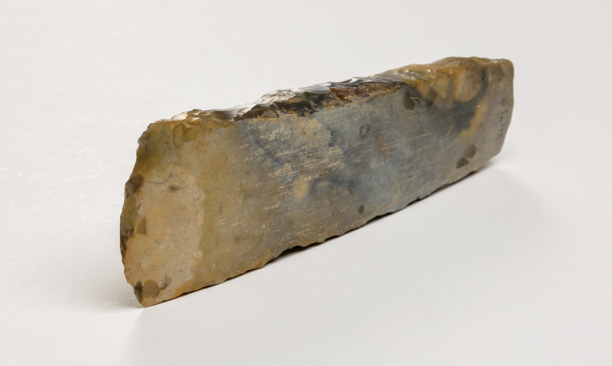 Flintyxa av brungrå flinta, närmast tunnackig, slipade bredsidor med avrundad egg. Rektangulär genomskärning.