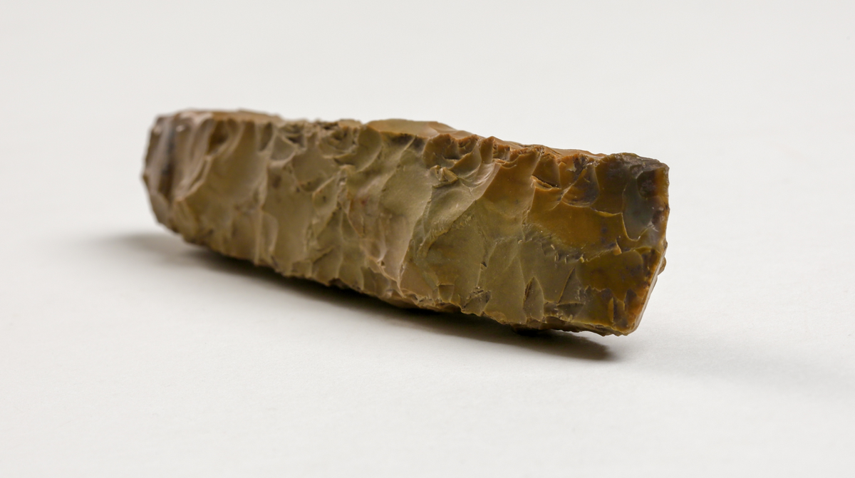 Rätmejsel av brun flinta, smal, tjocknackig, oslipad med svagt avrundad egg. Kvadratisk genomskärning.