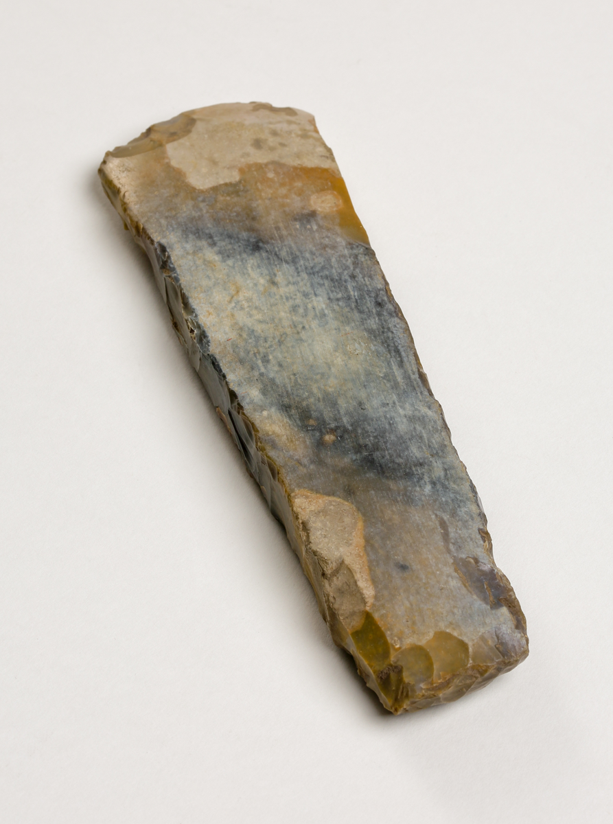 Flintyxa av brungrå flinta, närmast tunnackig, slipade bredsidor med avrundad egg. Rektangulär genomskärning.