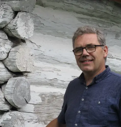 En mann med blå skjorte og briller står ved et laftet hushjørne som er hvitkalket.