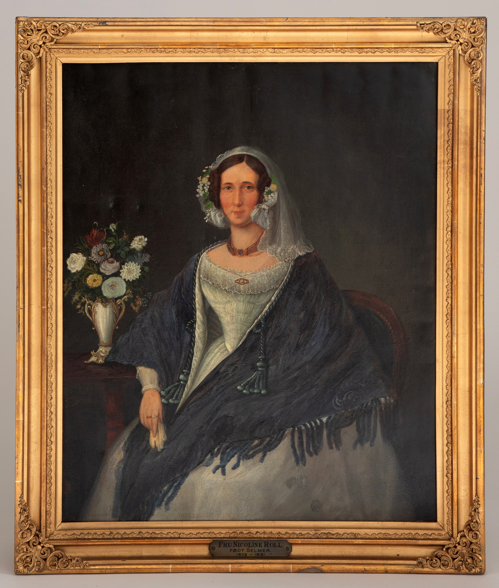 Portrett av en sittende kvinne i trekvart-figur. Kvinnen er iført en hvit kjole og et stort sjal i nyanser av blått og svart. Hun har et hvit slør med kniplinger over hodet, samt hodepynt av hvite blomster med grønne blad. På et brunt bord står det en hvit vase med hvite, røde og blå blomster. Bakgrunnen er mørk.