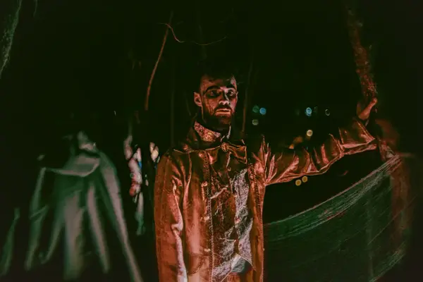 Foto av en mann i kostyme og skummel sminke som står i en mørk hage. 