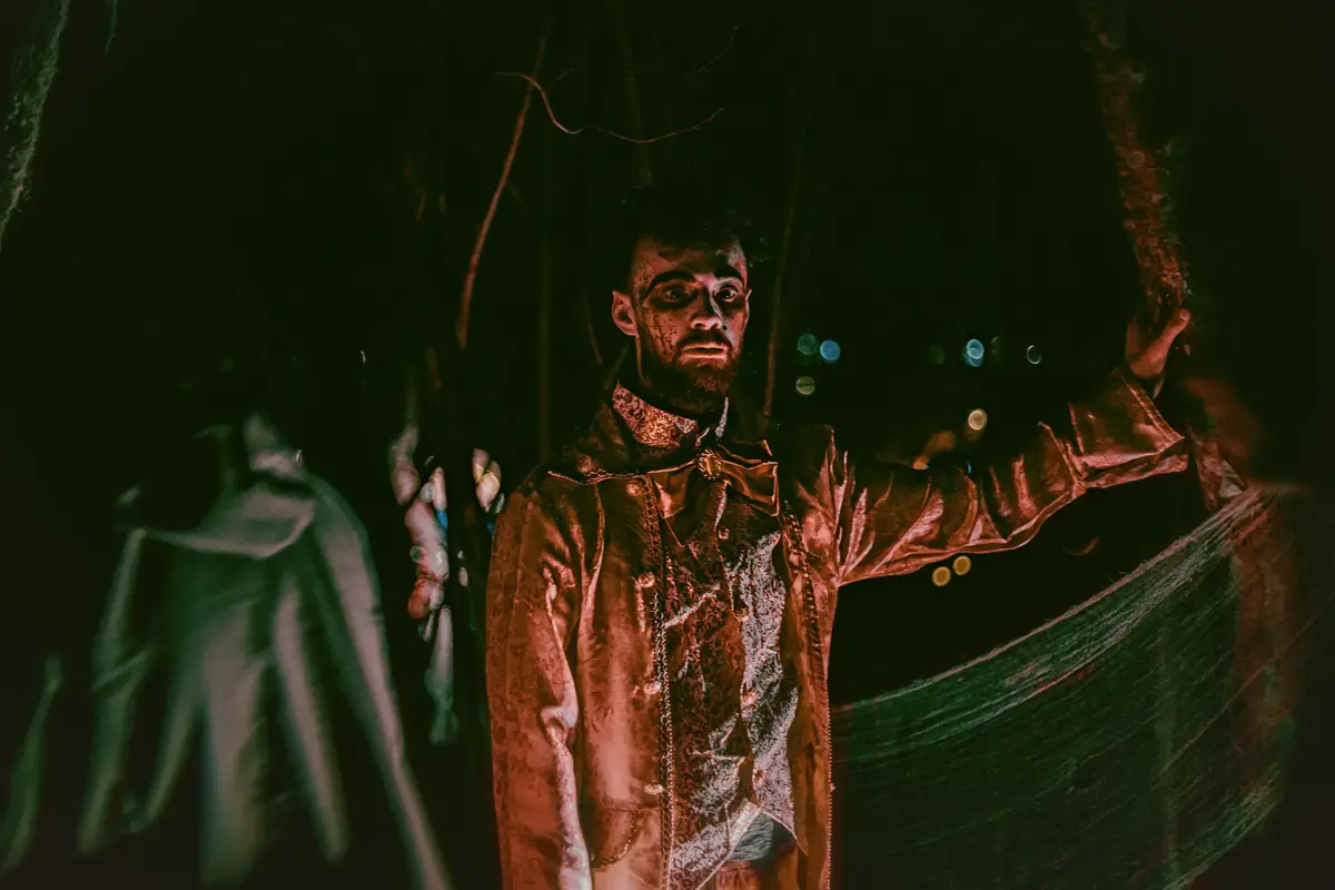 Foto av en mann i kostyme og skummel sminke som står i en mørk hage. 