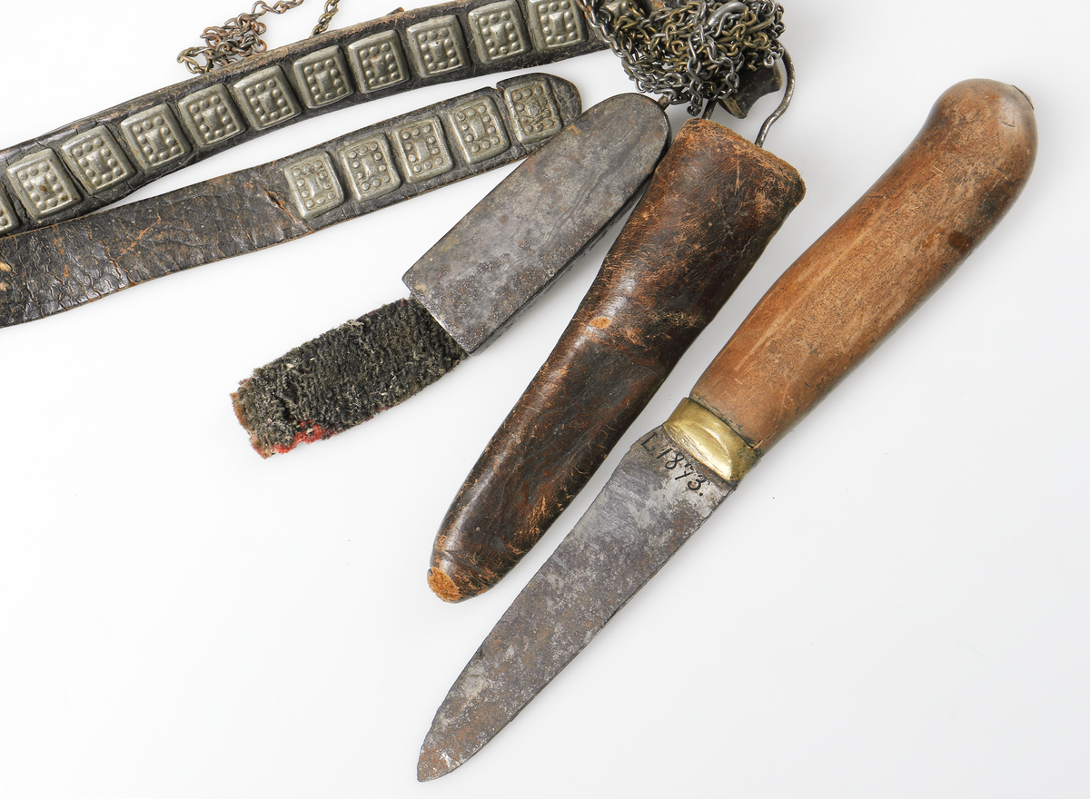 Så kallat kvinnobälte med kniv och nålhus från Dalarna. Bältet är gjort av läder och är prytt med små gjutna tennplattor. Nålhus och kniv är av järn, knivskaftet tillverkat av päronträ med mässingsdetalj ned mot knivbladet.