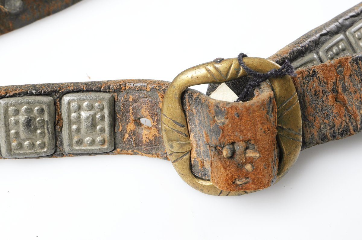 Så kallat kvinnobälte med kniv och nålhus från Dalarna. Bältet är gjort av läder och är prytt med små gjutna tennplattor. Nålhus och kniv är av järn, knivskaftet tillverkat av päronträ med mässingsdetalj ned mot knivbladet.