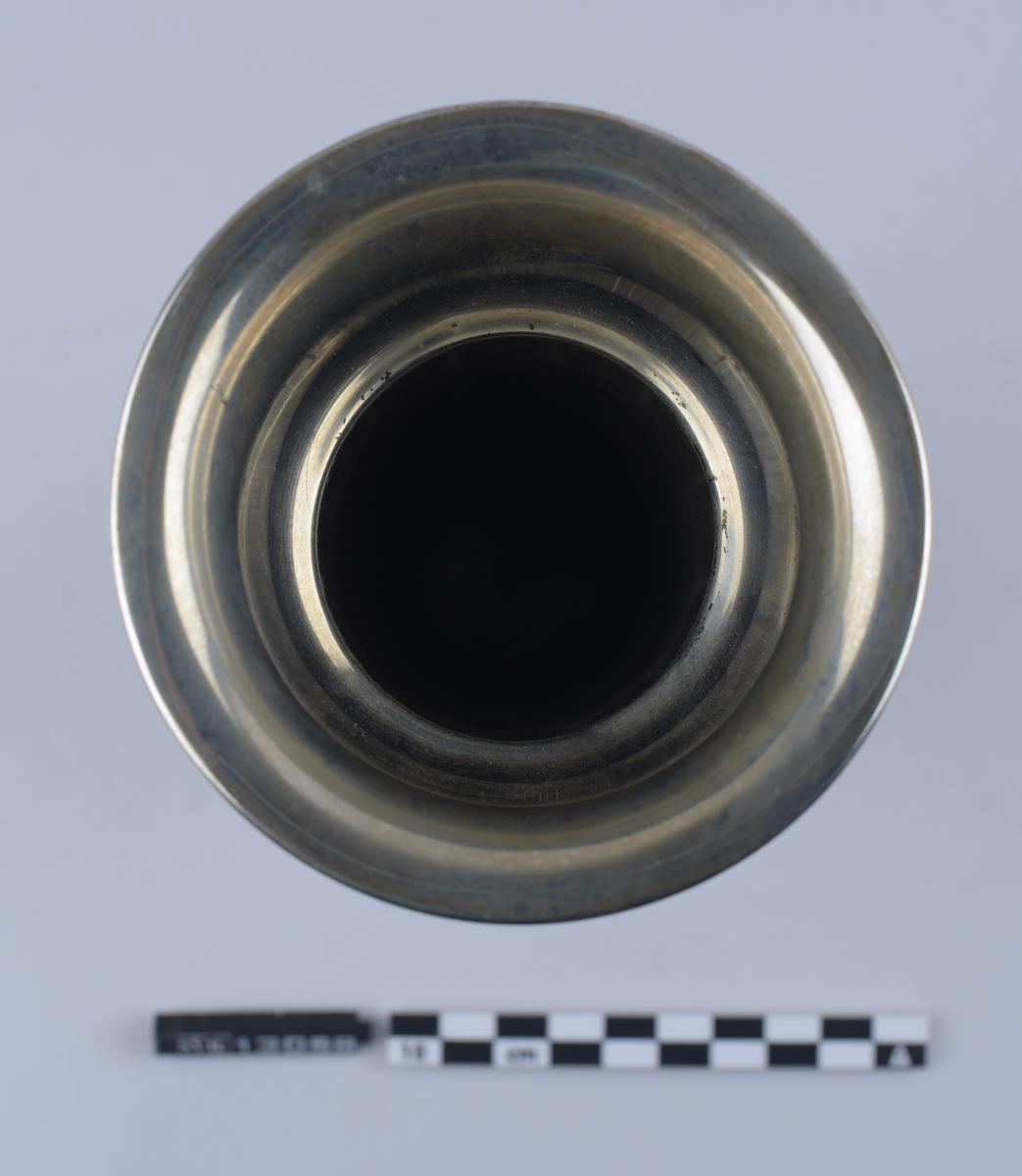 Metallgjenstand, sølvfarget, står på en sirkulær base, har en tilnærmet rund kropp og en sirkulær, utflatende topp. Hul inni. Sidene er avflatet og delt inn i 12 segment. Toppen er bredere enn basen. Øvre kant strekker seg 1 centimeter ut fra gjenstandens kropp og danner en buet lippe.