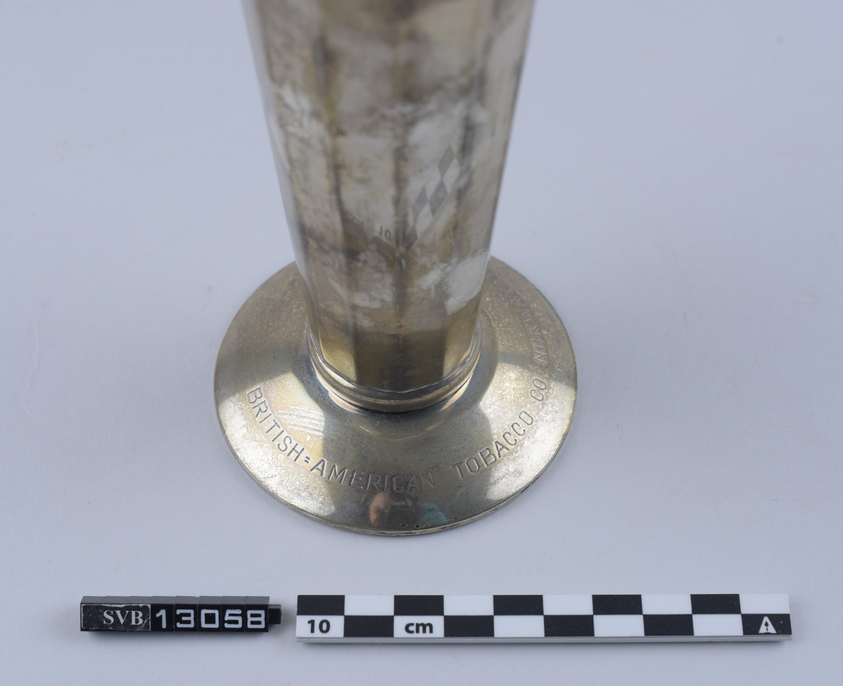 Metallgjenstand, sølvfarget, står på en sirkulær base, har en tilnærmet rund kropp og en sirkulær, utflatende topp. Hul inni. Sidene er avflatet og delt inn i 12 segment. Toppen er bredere enn basen. Øvre kant strekker seg 1 centimeter ut fra gjenstandens kropp og danner en buet lippe.