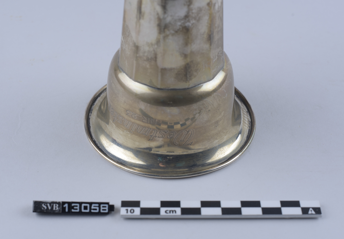 Metallgjenstand, sølvfarget, står på en sirkulær base, har en tilnærmet rund kropp og en sirkulær, utflatende topp. Hul inni. Sidene er avflatet og delt inn i 12 segment. Toppen er bredere enn basen. Øvre kant strekker seg 1 centimeter ut fra gjenstandens kropp og danner en buet lippe.