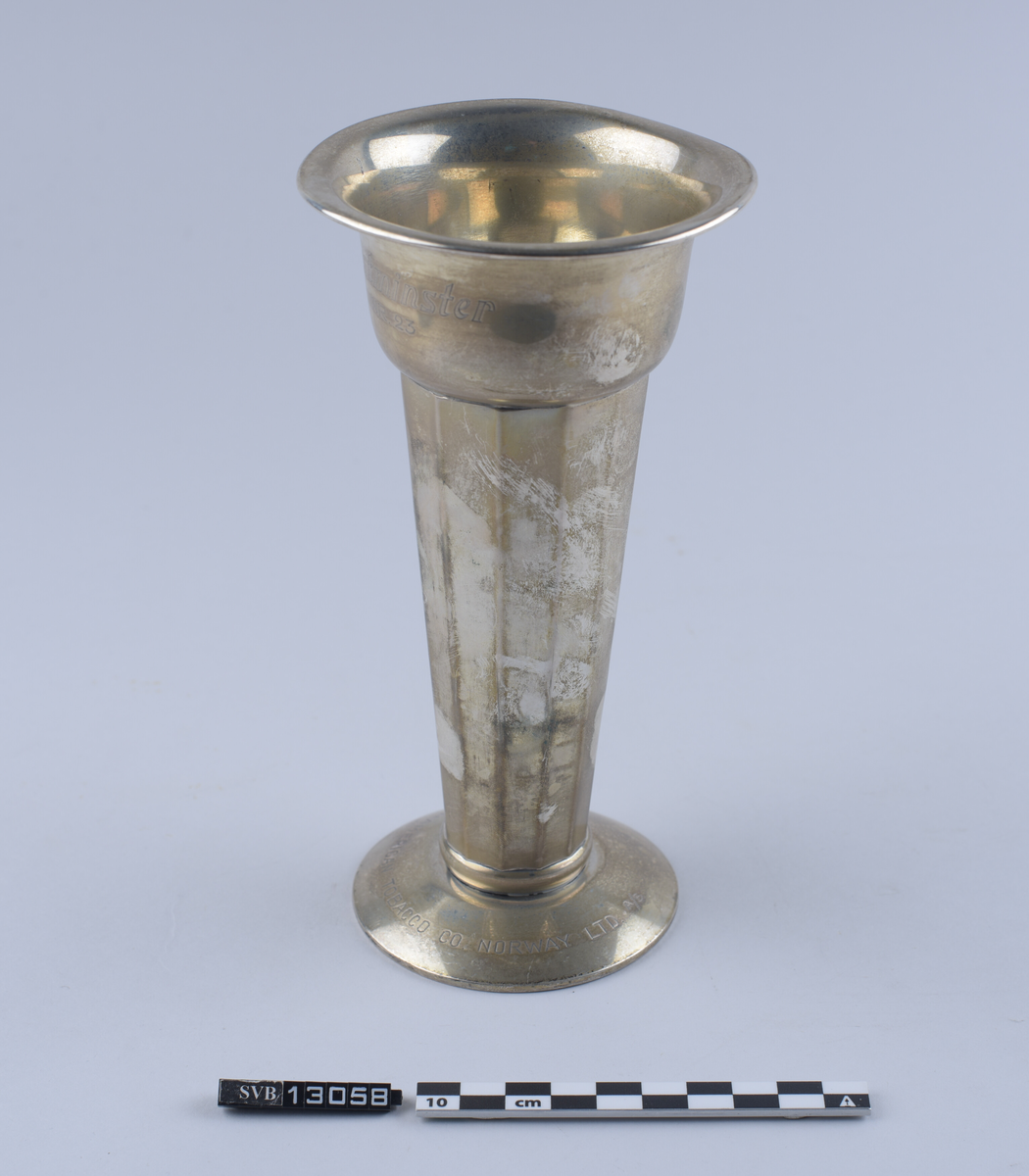 Metallgjenstand, sølvfarget, står på en sirkulær base, har en tilnærmet rund kropp og en sirkulær, utflatende topp. Hul inni. Sidene er avflatet og delt inn i 12 segment. Toppen er bredere enn basen. Øvre kant strekker seg 1 centimeter ut fra gjenstandens kropp og danner en buet lippe.