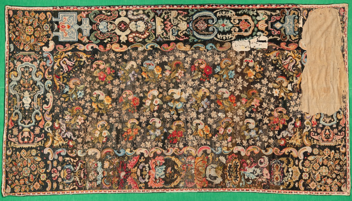 Linbotten med broderier i framför allt ull. Vissa detaljer troligen gjorda i silke. Lagningarna utförda i ull och bomull. Broderiet gjort i korsstygn. De äldsta delarna (bården) är gjorda i olika stora stygn varför ytans karaktär varierar. Mönster som påminner om pompeijansk stil med bland annat kartusher, urnor och växtornament. Mittpartiet inramat med roccaier eller akantusblad, blombotten med liljor och rosor samt vinrankor. Längst ena långsidan även inslag av korall. Hörn med spiralformade blomstergirlanger, dessa med drag av folkligt broderi.
Brunsvart botten, ojämt blekt. Merparten beigegröna nyanser på växtornamenten. Blåa och röda inslag där vissa röda delar skulle kunna vara anilinfärg, troligen i övrigt växtfärg.