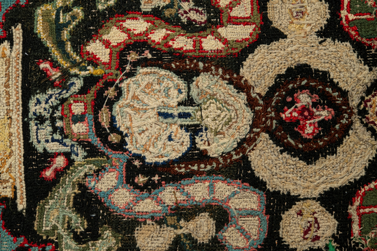 Linbotten med broderier i framför allt ull. Vissa detaljer troligen gjorda i silke. Lagningarna utförda i ull och bomull. Broderiet gjort i korsstygn. De äldsta delarna (bården) är gjorda i olika stora stygn varför ytans karaktär varierar. Mönster som påminner om pompeijansk stil med bland annat kartusher, urnor och växtornament. Mittpartiet inramat med roccaier eller akantusblad, blombotten med liljor och rosor samt vinrankor. Längst ena långsidan även inslag av korall. Hörn med spiralformade blomstergirlanger, dessa med drag av folkligt broderi.
Brunsvart botten, ojämt blekt. Merparten beigegröna nyanser på växtornamenten. Blåa och röda inslag där vissa röda delar skulle kunna vara anilinfärg, troligen i övrigt växtfärg.