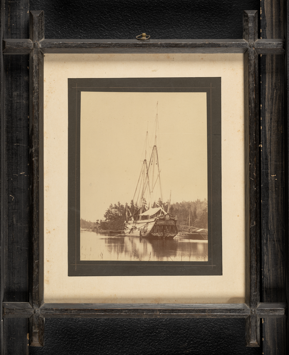 Ram av trä från kungliga skonerten Amphion.
Tryckt textskylt på tavlans baksida:
"Historien om ett kungsfartyg.
Amphion byggdes år 1778 på Djurgårdsvarfvet och förärades det derpå följande året Stockholms borgerskap åt Gustaf III för att begagnas vid de lustresor som konungen vid denna tid ofta företog. Men först 10 år senare, 1788, började Amphions egentliga historia. Kriget med Ryssland hade då utbrutit. Jemte galérflottan gick Gustaf III på Amphion öfver till Finland och landsteg i Helssingfors. Under hela det ryska fälttåget bodde konungen, med korta afbrott, om bord å skonerten. Här höllos alla rådplägningarna under den bekymmersamma tiden före Anjalaförbundets sprängning. Om konungen på Amphion gjorde öferfarten från Åbo till Stockholm i slutet av 1788 vet man ej med visshet; troligt är dock, att så var förhållandet.

Då Amphion i mars 1790 för tredje gången seglade övfer till Finland, låg ännu isen qvar i Östersjön, så att man måste såga väg för skonerten. På Ålands haf var man utsatt för en svår storm,så att nästan alla om bord varande, till och med sjövanan matroser, blevo sjösjuka, "Konungen låg i sin säng och luktade på färska citroner och apelsiner, hvarigenom han undvek den allmänna olägenheten." 

Den 3 juli skedde sedan den bekanta genombrytningen vid Aspö, det s.k. Wiborgska gatloppet. Amphion låg under de närmaste föregående dagarne på Wiborgs redd. Den 2 på aftonen gick konungen från skonerten och steg om bord på chefsfartyget Kung Gustaf III. Det var hans mening att genast begifva sig till baka till sitt eget fartyg, men genom ryska flottans operationer hindrades han derifrån och steg först några dagar senare (den 5) i Svensksund åter ombord på Amphion.

Under genombrytningen befann sig Amphion jemte jakten Esplendian i midten af svanska skärgårdsflottans lilnie. Amphion med sina prydnader och förgyllningar ådrog sig naturligtvis fiendens uppmärksamhet och blef genast en skottafla; men blott rorgängaren blef dödsskjuten: en kula slet bort magen på honom. Äfven Amphions aktermast bortsköts nästan helt och hållet. Sjelf deltog skonerten ej i striden, då den endast var bestyckad med några salutnickor.

Då Amphion sedan vid mörkrets inbrott skulle passera Aspöskären, råkade han i kollision med åtskilliga andra fartyg, hvilkas rår fastnade i Amphions sönderskjutna tackling, som måste än vidare sönderhuggas. På andra sidan om den klippa, vid hvilken Amphion var förankrad, låg en mängd ryska fartyg, hvilka dock i brist på ammunition ej kunde upptaga någon strid med fienden.

På morgonen lyckdes det icke att få Amphion under segel på grund af det trånga farvattnet. Det tycktes icke återstå annat än att bränna skonerten för att ej låta honom falla i händerna på fiendern, som gjorde min af att gå anfallsvis till väga. Men genom befälhavarens, löjtnant Eskilssons, rådighet fick man slutligen upp seglen.

Då skonerten sedan utkommit i Öppna sjön, passerade den helt nära en del af den fiendtliga eskadern, som under stormen föregående natt blifvit illa medfaren och som nu på grund af Amphionas höga tackling trodde den var fregatt, hvarför ryssarne gjorde sig i ordning till försver. Amphion afsköt sina saluterkanoner mot fienderna och fortsatte sin färd. Ännu vori ej alla svårigheter öfvervunna, löjtnant Eskilsons hele mod och skicklighet sattes ännu allt jemt på prof, men slutligen kom han med kungsfartyget så långt från fienden, att all fara var öfver. Denne löjtnant Eskilson blef sedemera under förmyndareregeringen för någon mindre förseelse dömd att arkubuseras, men "benådades med lifstids fängelse" och transporterades först till Carlstens och sedan till Waxholms fästning, der han dog i vansinne, troligen till följd av den svåra behandlingen.

Den 8 juni 1790 höll konungen på Amphion den konselj, der det andra slaget vid Svensksund beslöts. Tidigt på morgonen lemnade konungen sitt fartyg för att intaga sin plats på chefsskeppet. Amphion deltog ej aktift i striden. Under densamma lät general Klingspor, som ej tycktes hafva hyst stora förhoppningar om seger, från mphion föra i land konungens silfver, garderob, kokkärl, ja till och med de väggfasta speglarne för att skonerten hvilket ögonblick som helst skulle kunna brännas och undgå att råka i fiendens händer. Konungen var mycket missnöjd häröfver, ty då han efter det vunne slaget begaf sig ombord på sitt fartyg, hade han icke funnit en säng att ligga i, icke en bit mat att äta.

Te Deum öfver den vunna segern hölls den 22 i samma månad på drottningens namnsdag. Gudtjemsten försiggick på en holme, framför hvilken hela svenska flottan låg förankrad. Efter ceremonins slut begav sig konungen till Amphion, som åter stälts i ordning, och det var anrättad en festmåltid för 50 personer under ett på däcket uppslaget tält. Före festen utdelade konungen åtskilliga belöningar i form af medaljer och ordnar.

Här en liten anekdot.
Öfverste Stedingk, som slagits tappert under bataljen, blef högligen uppretad öfver att öfverste Cronstedt framför honom erhållit stora korset af svärdorden, men han sjelf blott en hedervärja. Vid middagsbordet kunde han ej återhålla sin harm, ehuru konungen ej låtsades märka det. Man berättade sedan att han, för att löjliggöra en del af de gifna belöningarna, med stora ceremonier låtit framföra en på hans fartyg befintlig get, som under fälttåget försett honom med mjlök, för att efter ett långt, högtidligt tal om hennes förtjenster förklara att hon blifvit upphöjd till rang, heder och värdighet at ko, dock med bibehållande af sin förra utfodring.

Den 14 augusti slöts freden i Wärälä, och den 17 på eftermiggaden samlades alla de svenska officerarne om bord å Amphion, der konungen tog afsked af dem. Den 28 anlände han med skonerten till Stockholm."