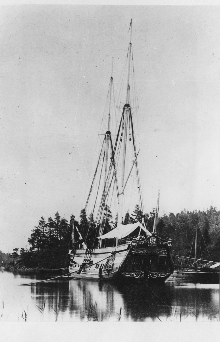 Ram av trä från kungliga skonerten Amphion.
Tryckt textskylt på tavlans baksida:
"Historien om ett kungsfartyg.
Amphion byggdes år 1778 på Djurgårdsvarfvet och förärades det derpå följande året Stockholms borgerskap åt Gustaf III för att begagnas vid de lustresor som konungen vid denna tid ofta företog. Men först 10 år senare, 1788, började Amphions egentliga historia. Kriget med Ryssland hade då utbrutit. Jemte galérflottan gick Gustaf III på Amphion öfver till Finland och landsteg i Helssingfors. Under hela det ryska fälttåget bodde konungen, med korta afbrott, om bord å skonerten. Här höllos alla rådplägningarna under den bekymmersamma tiden före Anjalaförbundets sprängning. Om konungen på Amphion gjorde öferfarten från Åbo till Stockholm i slutet av 1788 vet man ej med visshet; troligt är dock, att så var förhållandet.

Då Amphion i mars 1790 för tredje gången seglade övfer till Finland, låg ännu isen qvar i Östersjön, så att man måste såga väg för skonerten. På Ålands haf var man utsatt för en svår storm,så att nästan alla om bord varande, till och med sjövanan matroser, blevo sjösjuka, "Konungen låg i sin säng och luktade på färska citroner och apelsiner, hvarigenom han undvek den allmänna olägenheten." 

Den 3 juli skedde sedan den bekanta genombrytningen vid Aspö, det s.k. Wiborgska gatloppet. Amphion låg under de närmaste föregående dagarne på Wiborgs redd. Den 2 på aftonen gick konungen från skonerten och steg om bord på chefsfartyget Kung Gustaf III. Det var hans mening att genast begifva sig till baka till sitt eget fartyg, men genom ryska flottans operationer hindrades han derifrån och steg först några dagar senare (den 5) i Svensksund åter ombord på Amphion.

Under genombrytningen befann sig Amphion jemte jakten Esplendian i midten af svanska skärgårdsflottans lilnie. Amphion med sina prydnader och förgyllningar ådrog sig naturligtvis fiendens uppmärksamhet och blef genast en skottafla; men blott rorgängaren blef dödsskjuten: en kula slet bort magen på honom. Äfven Amphions aktermast bortsköts nästan helt och hållet. Sjelf deltog skonerten ej i striden, då den endast var bestyckad med några salutnickor.

Då Amphion sedan vid mörkrets inbrott skulle passera Aspöskären, råkade han i kollision med åtskilliga andra fartyg, hvilkas rår fastnade i Amphions sönderskjutna tackling, som måste än vidare sönderhuggas. På andra sidan om den klippa, vid hvilken Amphion var förankrad, låg en mängd ryska fartyg, hvilka dock i brist på ammunition ej kunde upptaga någon strid med fienden.

På morgonen lyckdes det icke att få Amphion under segel på grund af det trånga farvattnet. Det tycktes icke återstå annat än att bränna skonerten för att ej låta honom falla i händerna på fiendern, som gjorde min af att gå anfallsvis till väga. Men genom befälhavarens, löjtnant Eskilssons, rådighet fick man slutligen upp seglen.

Då skonerten sedan utkommit i Öppna sjön, passerade den helt nära en del af den fiendtliga eskadern, som under stormen föregående natt blifvit illa medfaren och som nu på grund af Amphionas höga tackling trodde den var fregatt, hvarför ryssarne gjorde sig i ordning till försver. Amphion afsköt sina saluterkanoner mot fienderna och fortsatte sin färd. Ännu vori ej alla svårigheter öfvervunna, löjtnant Eskilsons hele mod och skicklighet sattes ännu allt jemt på prof, men slutligen kom han med kungsfartyget så långt från fienden, att all fara var öfver. Denne löjtnant Eskilson blef sedemera under förmyndareregeringen för någon mindre förseelse dömd att arkubuseras, men "benådades med lifstids fängelse" och transporterades först till Carlstens och sedan till Waxholms fästning, der han dog i vansinne, troligen till följd av den svåra behandlingen.

Den 8 juni 1790 höll konungen på Amphion den konselj, der det andra slaget vid Svensksund beslöts. Tidigt på morgonen lemnade konungen sitt fartyg för att intaga sin plats på chefsskeppet. Amphion deltog ej aktift i striden. Under densamma lät general Klingspor, som ej tycktes hafva hyst stora förhoppningar om seger, från mphion föra i land konungens silfver, garderob, kokkärl, ja till och med de väggfasta speglarne för att skonerten hvilket ögonblick som helst skulle kunna brännas och undgå att råka i fiendens händer. Konungen var mycket missnöjd häröfver, ty då han efter det vunne slaget begaf sig ombord på sitt fartyg, hade han icke funnit en säng att ligga i, icke en bit mat att äta.

Te Deum öfver den vunna segern hölls den 22 i samma månad på drottningens namnsdag. Gudtjemsten försiggick på en holme, framför hvilken hela svenska flottan låg förankrad. Efter ceremonins slut begav sig konungen till Amphion, som åter stälts i ordning, och det var anrättad en festmåltid för 50 personer under ett på däcket uppslaget tält. Före festen utdelade konungen åtskilliga belöningar i form af medaljer och ordnar.

Här en liten anekdot.
Öfverste Stedingk, som slagits tappert under bataljen, blef högligen uppretad öfver att öfverste Cronstedt framför honom erhållit stora korset af svärdorden, men han sjelf blott en hedervärja. Vid middagsbordet kunde han ej återhålla sin harm, ehuru konungen ej låtsades märka det. Man berättade sedan att han, för att löjliggöra en del af de gifna belöningarna, med stora ceremonier låtit framföra en på hans fartyg befintlig get, som under fälttåget försett honom med mjlök, för att efter ett långt, högtidligt tal om hennes förtjenster förklara att hon blifvit upphöjd till rang, heder och värdighet at ko, dock med bibehållande af sin förra utfodring.

Den 14 augusti slöts freden i Wärälä, och den 17 på eftermiggaden samlades alla de svenska officerarne om bord å Amphion, der konungen tog afsked af dem. Den 28 anlände han med skonerten till Stockholm."