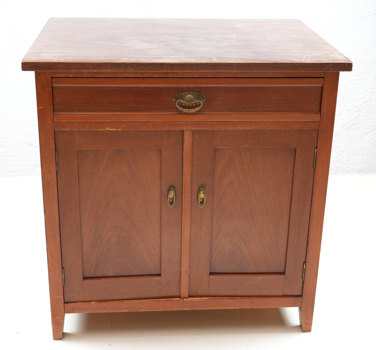 Brunbeisa kommode i heiltre. Ei skuffe øverst med messinghandtak. Under skuffa er det eit skap med doble skapdører og messinghandtak. Enkel profil på skapdørene. Ei hylleplate inni av heiltre. Kommoden står på fire bein. 