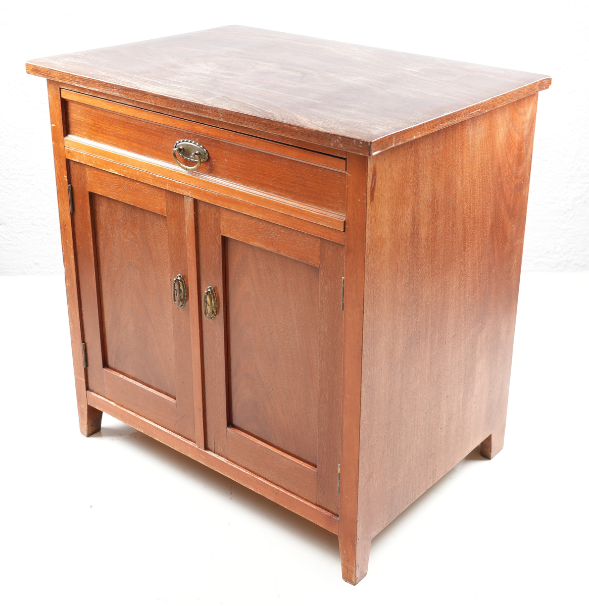 Brunbeisa kommode i heiltre. Ei skuffe øverst med messinghandtak. Under skuffa er det eit skap med doble skapdører og messinghandtak. Enkel profil på skapdørene. Ei hylleplate inni av heiltre. Kommoden står på fire bein. 