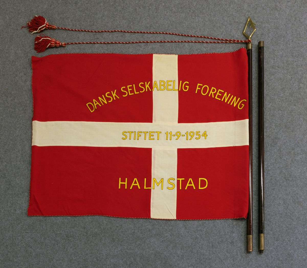 AHF000806a Dansk flagga, dubbel fanduk i rött och vitt, tuskaftat ylle. Kantad med röd och vit yllesnodd. Maskinbroderad text i konstsilke. 
AHF000806b Tvådelad stång med fantopp, snodd med tofsar i rött och vitt silke med träkärna. 
AHF000806c Bärrem av brunt läder. 
AHF000806d Fodral.