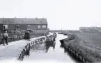 Foto av de traditionella nederländska fiskebyarna där kanalen kantas av byggnader och en gångväg där människor syns. I vattnet ligger det små båtar och två kvinnor för fram en båt, 1924-25.