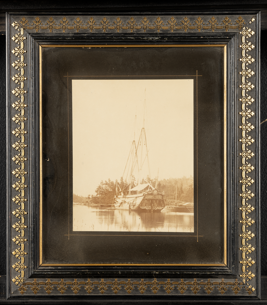 Svart ram med gulddekor.

Tryckt textskylt på tavlans baksida:
"Historien om ett kungsfartyg.
Amphion byggdes år 1778 på Djurgårdsvarfvet och förärades det derpå följande året Stockholms borgerskap åt Gustaf III för att begagnas vid de lustresor som konungen vid denna tid ofta företog. Men först 10 år senare, 1788, började Amphions egentliga historia. Kriget med Ryssland hade då utbrutit. Jemte galérflottan gick Gustaf III på Amphion öfver till Finland och landsteg i Helssingfors. Under hela det ryska fälttåget bodde konungen, med korta afbrott, om bord å skonerten. Här höllos alla rådplägningarna under den bekymmersamma tiden före Anjalaförbundets sprängning. Om konungen på Amphion gjorde öferfarten från Åbo till Stockholm i slutet av 1788 vet man ej med visshet; troligt är dock, att så var förhållandet.

Då Amphion i mars 1790 för tredje gången seglade övfer till Finland, låg ännu isen qvar i Östersjön, så att man måste såga väg för skonerten. På Ålands haf var man utsatt för en svår storm,så att nästan alla om bord varande, till och med sjövanan matroser, blevo sjösjuka, "Konungen låg i sin säng och luktade på färska citroner och apelsiner, hvarigenom han undvek den allmänna olägenheten." 

Den 3 juli skedde sedan den bekanta genombrytningen vid Aspö, det s.k. Wiborgska gatloppet. Amphion låg under de närmaste föregående dagarne på Wiborgs redd. Den 2 på aftonen gick konungen från skonerten och steg om bord på chefsfartyget Kung Gustaf III. Det var hans mening att genast begifva sig till baka till sitt eget fartyg, men genom ryska flottans operationer hindrades han derifrån och steg först några dagar senare (den 5) i Svensksund åter ombord på Amphion.

Under genombrytningen befann sig Amphion jemte jakten Esplendian i midten af svanska skärgårdsflottans lilnie. Amphion med sina prydnader och förgyllningar ådrog sig naturligtvis fiendens uppmärksamhet och blef genast en skottafla; men blott rorgängaren blef dödsskjuten: en kula slet bort magen på honom. Äfven Amphions aktermast bortsköts nästan helt och hållet. Sjelf deltog skonerten ej i striden, då den endast var bestyckad med några salutnickor.

Då Amphion sedan vid mörkrets inbrott skulle passera Aspöskären, råkade han i kollision med åtskilliga andra fartyg, hvilkas rår fastnade i Amphions sönderskjutna tackling, som måste än vidare sönderhuggas. På andra sidan om den klippa, vid hvilken Amphion var förankrad, låg en mängd ryska fartyg, hvilka dock i brist på ammunition ej kunde upptaga någon strid med fienden.

På morgonen lyckdes det icke att få Amphion under segel på grund af det trånga farvattnet. Det tycktes icke återstå annat än att bränna skonerten för att ej låta honom falla i händerna på fiendern, som gjorde min af att gå anfallsvis till väga. Men genom befälhavarens, löjtnant Eskilssons, rådighet fick man slutligen upp seglen.

Då skonerten sedan utkommit i Öppna sjön, passerade den helt nära en del af den fiendtliga eskadern, som under stormen föregående natt blifvit illa medfaren och som nu på grund af Amphionas höga tackling trodde den var fregatt, hvarför ryssarne gjorde sig i ordning till försver. Amphion afsköt sina saluterkanoner mot fienderna och fortsatte sin färd. Ännu vori ej alla svårigheter öfvervunna, löjtnant Eskilsons hele mod och skicklighet sattes ännu allt jemt på prof, men slutligen kom han med kungsfartyget så långt från fienden, att all fara var öfver. Denne löjtnant Eskilson blef sedemera under förmyndareregeringen för någon mindre förseelse dömd att arkubuseras, men "benådades med lifstids fängelse" och transporterades först till Carlstens och sedan till Waxholms fästning, der han dog i vansinne, troligen till följd av den svåra behandlingen.

Den 8 juni 1790 höll konungen på Amphion den konselj, der det andra slaget vid Svensksund beslöts. Tidigt på morgonen lemnade konungen sitt fartyg för att intaga sin plats på chefsskeppet. Amphion deltog ej aktift i striden. Under densamma lät general Klingspor, som ej tycktes hafva hyst stora förhoppningar om seger, från mphion föra i land konungens silfver, garderob, kokkärl, ja till och med de väggfasta speglarne för att skonerten hvilket ögonblick som helst skulle kunna brännas och undgå att råka i fiendens händer. Konungen var mycket missnöjd häröfver, ty då han efter det vunne slaget begaf sig ombord på sitt fartyg, hade han icke funnit en säng att ligga i, icke en bit mat att äta.

Te Deum öfver den vunna segern hölls den 22 i samma månad på drottningens namnsdag. Gudtjemsten försiggick på en holme, framför hvilken hela svenska flottan låg förankrad. Efter ceremonins slut begav sig konungen till Amphion, som åter stälts i ordning, och det var anrättad en festmåltid för 50 personer under ett på däcket uppslaget tält. Före festen utdelade konungen åtskilliga belöningar i form af medaljer och ordnar.

Här en liten anekdot.
Öfverste Stedingk, som slagits tappert under bataljen, blef högligen uppretad öfver att öfverste Cronstedt framför honom erhållit stora korset af svärdorden, men han sjelf blott en hedervärja. Vid middagsbordet kunde han ej återhålla sin harm, ehuru konungen ej låtsades märka det. Man berättade sedan att han, för att löjliggöra en del af de gifna belöningarna, med stora ceremonier låtit framföra en på hans fartyg befintlig get, som under fälttåget försett honom med mjlök, för att efter ett långt, högtidligt tal om hennes förtjenster förklara att hon blifvit upphöjd till rang, heder och värdighet at ko, dock med bibehållande af sin förra utfodring.

Den 14 augusti slöts freden i Wärälä, och den 17 på eftermiggaden samlades alla de svenska officerarne om bord å Amphion, der konungen tog afsked af dem. Den 28 anlände han med skonerten till Stockholm."