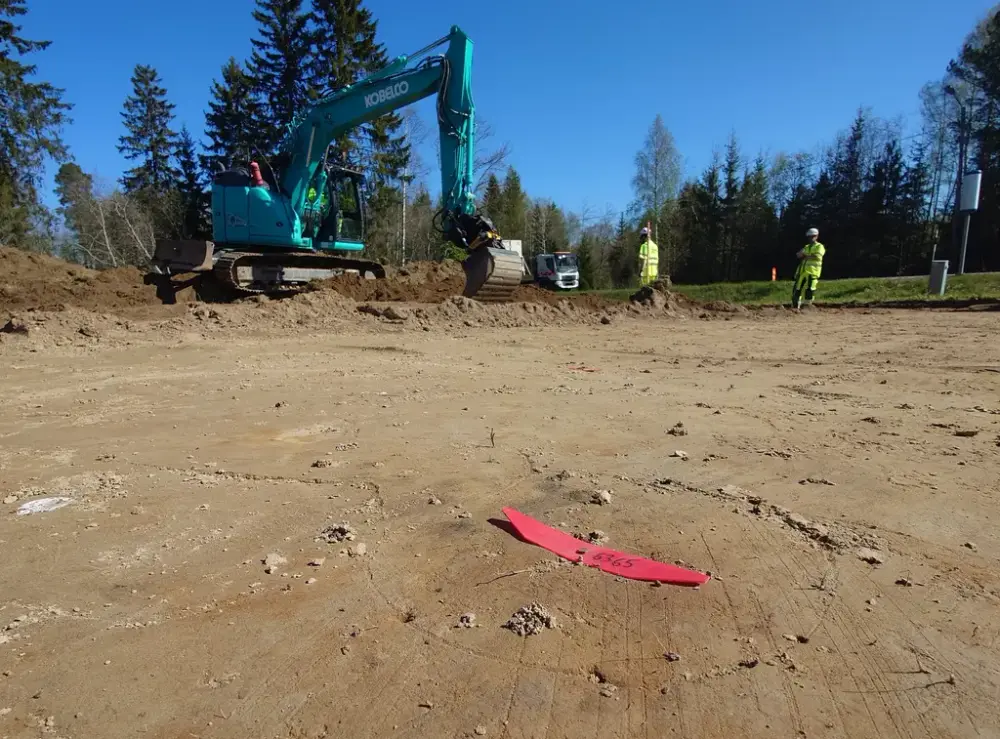 Arkeologisk utgrävning med två personer i varselkläder och en grävskopa i bakgrunden.