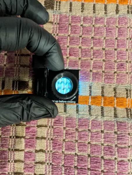 Händer med handskar håller ett förstoringsglas ovanför en väv.