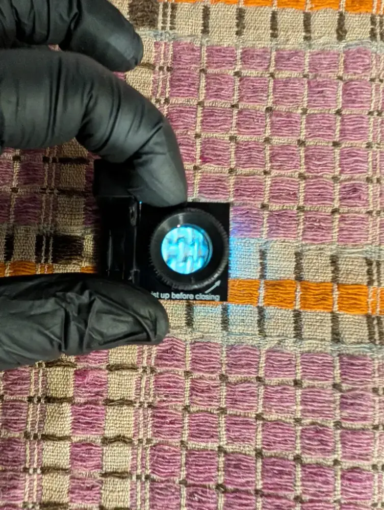 Händer med handskar håller ett förstoringsglas ovanför en väv.