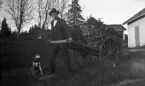 En ung man drar en kärra full med löv och grenar. En hund sitter bredvid honom på marken. I bakgrunden syns ett par uthus.