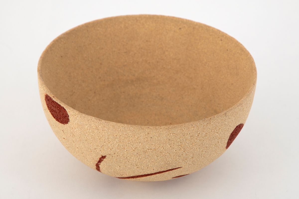 Rund skål i steingods. Halvkuleformet korpus på lav fotring. Skålen er uglasert og har en ru overflate. Dekorert med innlagte, abstrakte former i rødt.