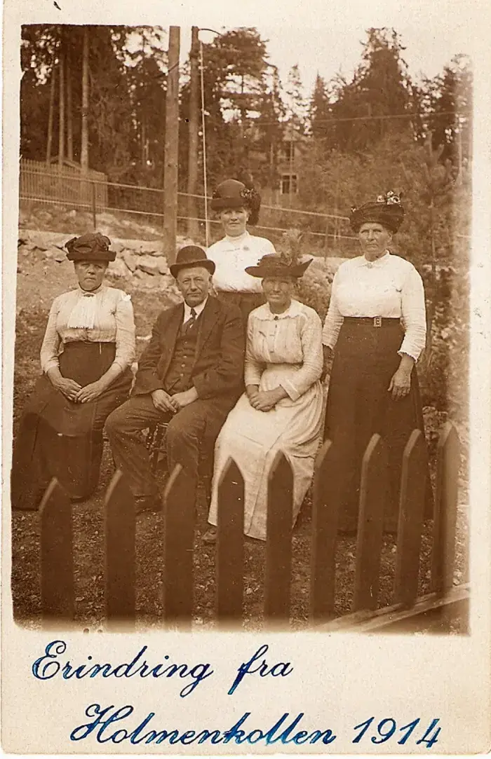 Historisk bilde av fire kvinner og en mann som sitter og står i en skråning bak et stakittgjerde. Påskrift: Erindring fra Holmenkollen 1914.